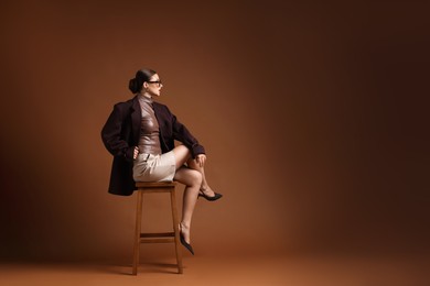 Photo of Young woman in stylish clothes on chair against brown background, space for text