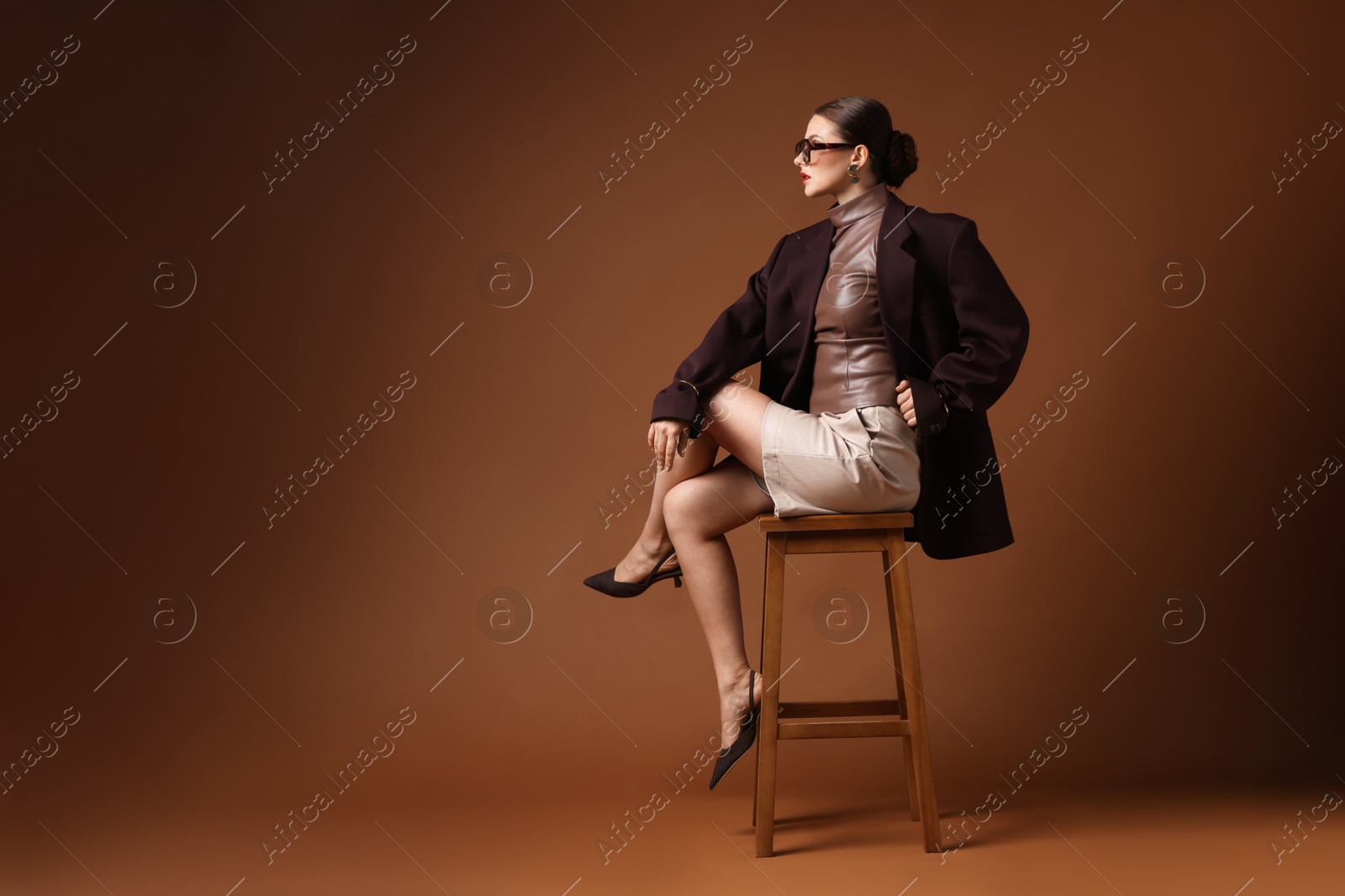 Young woman in stylish clothes on chair against brown background, space for text Photo of Young woman in stylish clothes on chair against brown background, space for text