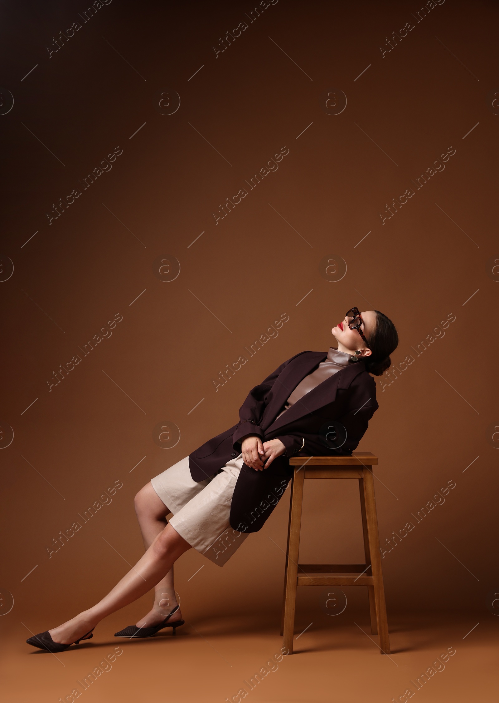 Young woman in stylish clothes on chair against brown background, space for text Photo of Young woman in stylish clothes on chair against brown background, space for text