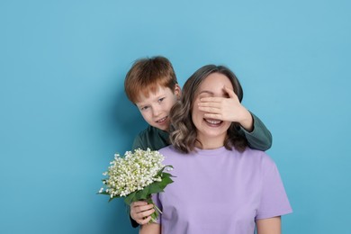 Photo of Boy surprising his mother with beautiful bouquet of lily-of-the-valley flowers on light blue background