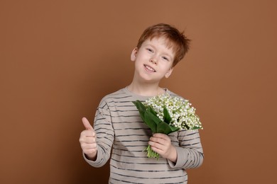 Photo of Boy with bouquet of beautiful lily-of-the-valley flowers showing thumbs up on brown background