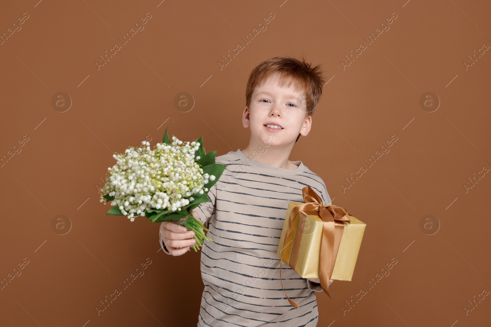 Boy with bouquet of beautiful lily-of-the-valley flowers and gift on brown background Photo of Boy with bouquet of beautiful lily-of-the-valley flowers and gift on brown background