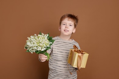 Photo of Boy with bouquet of beautiful lily-of-the-valley flowers and gift on brown background