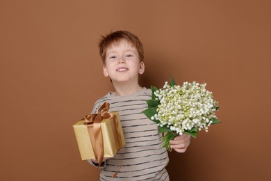 Photo of Boy with bouquet of beautiful lily-of-the-valley flowers and gift on brown background