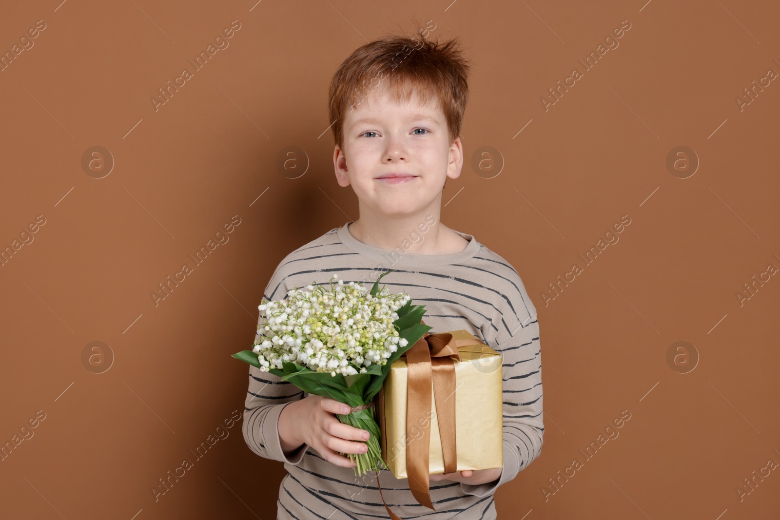 Boy with bouquet of beautiful lily-of-the-valley flowers and gift on brown background Photo of Boy with bouquet of beautiful lily-of-the-valley flowers and gift on brown background