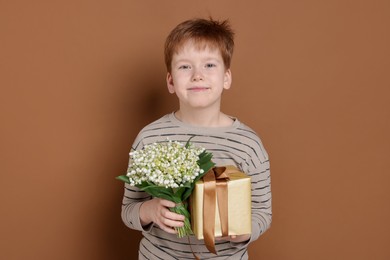Photo of Boy with bouquet of beautiful lily-of-the-valley flowers and gift on brown background