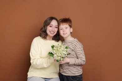Photo of Boy giving his mother beautiful bouquet of lily-of-the-valley flowers on brown background