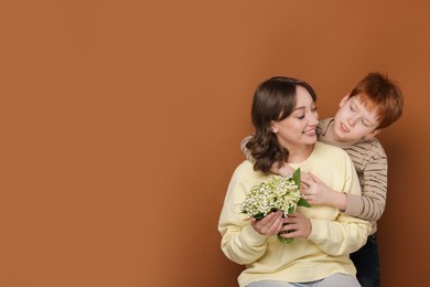 Photo of Boy giving his mother beautiful bouquet of lily-of-the-valley flowers on brown background. Space for text
