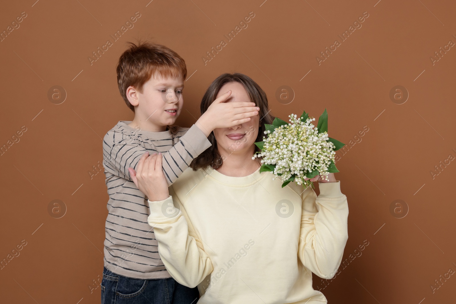 Boy surprising his mother with beautiful bouquet of lily-of-the-valley flowers on brown background Photo of Boy surprising his mother with beautiful bouquet of lily-of-the-valley flowers on brown background