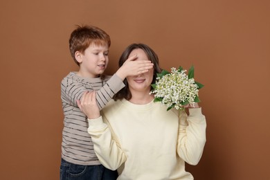 Photo of Boy surprising his mother with beautiful bouquet of lily-of-the-valley flowers on brown background