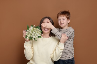 Photo of Boy surprising his mother with beautiful bouquet of lily-of-the-valley flowers on brown background