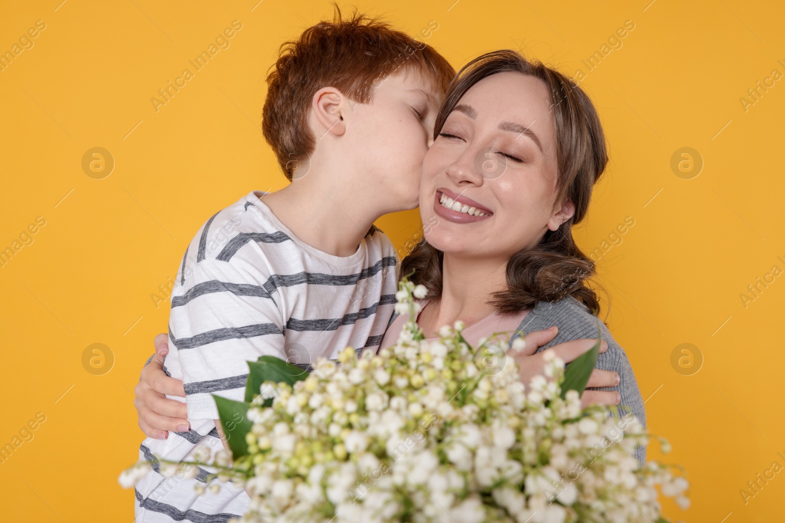 Boy kissing his mother and giving beautiful bouquet of lily-of-the-valley flowers on yellow background Photo of Boy kissing his mother and giving beautiful bouquet of lily-of-the-valley flowers on yellow background