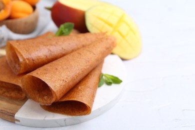 Photo of Delicious fruit leather rolls, mint, mango and apricots on white table, closeup. Space for text