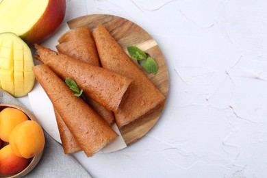 Photo of Delicious fruit leather rolls, mint, mango and apricots on white table, flat lay. Space for text