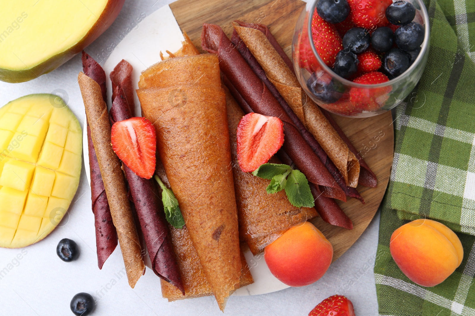 Delicious fruit leather rolls, mint, mango, apricots and berries on light table, flat lay Photo of Delicious fruit leather rolls, mint, mango, apricots and berries on light table, flat lay