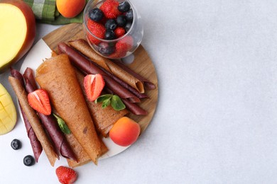 Photo of Delicious fruit leather rolls, mint, mango, apricots and berries on light table, flat lay. Space for text