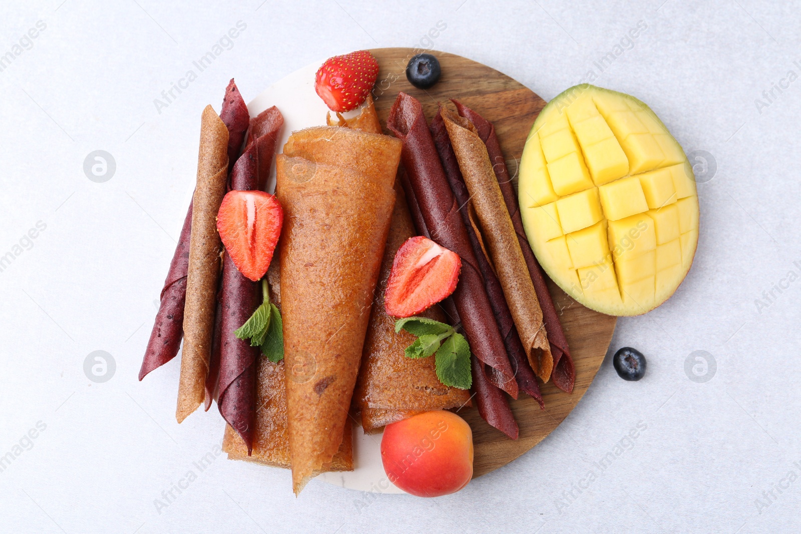 Delicious fruit leather rolls, mint, mango, apricot and berries on light table, flat lay Photo of Delicious fruit leather rolls, mint, mango, apricot and berries on light table, flat lay