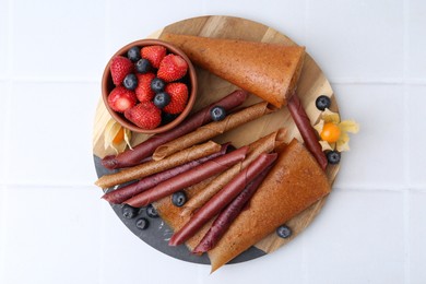 Photo of Delicious fruit leather rolls and berries on white tiled table, top view