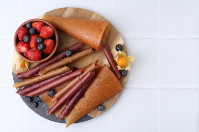 Photo of Delicious fruit leather rolls and berries on white tiled table, top view. Space for text