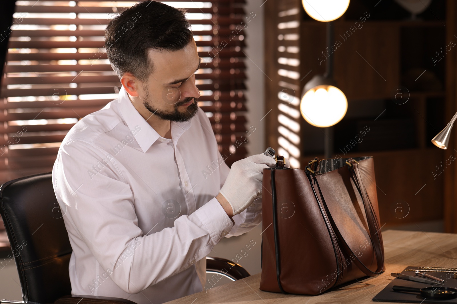 Appraiser with loupe evaluating leather bag at table indoors Photo of Appraiser with loupe evaluating leather bag at table indoors