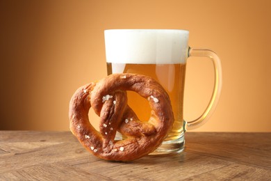 Photo of Tasty pretzel and glass mug of beer on wooden table against beige gradient background, closeup