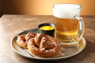 Photo of Tasty pretzels, sauce and glass mug beer on wooden table against beige gradient background, closeup