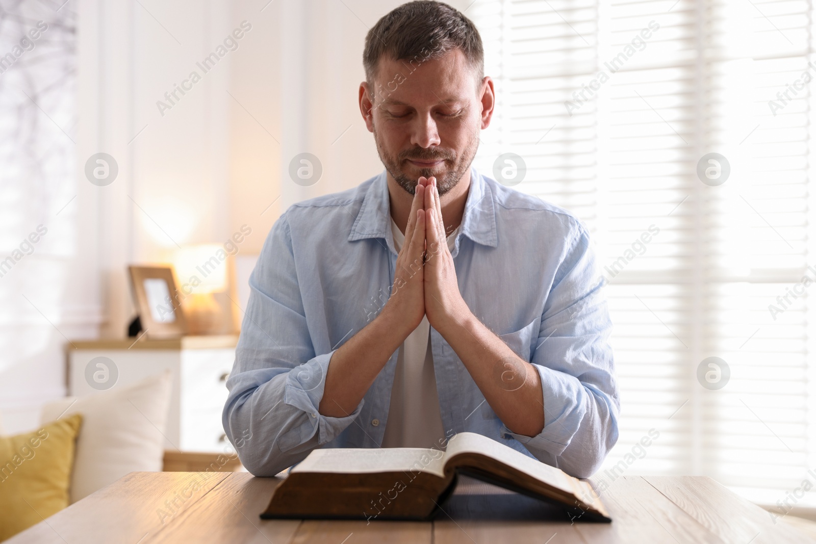 Man with Bible praying at table indoors Photo of Man with Bible praying at table indoors