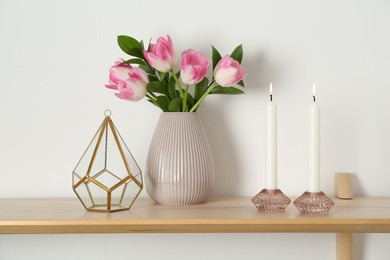 Photo of Elegant holders with burning candles, tulips in vase and other decor on wooden table near white wall