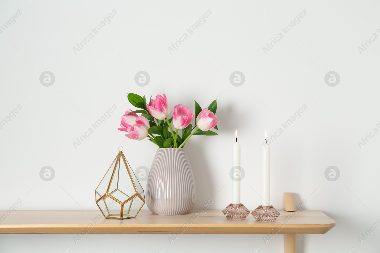 Elegant holders with burning candles, tulips in vase and other decor on wooden table near white wall Photo of Elegant holders with burning candles, tulips in vase and other decor on wooden table near white wall
