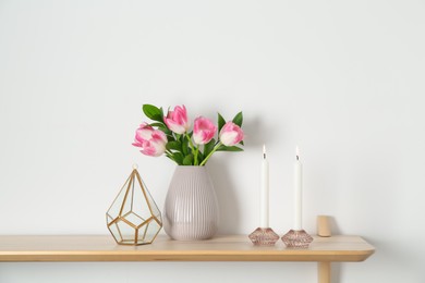 Photo of Elegant holders with burning candles, tulips in vase and other decor on wooden table near white wall