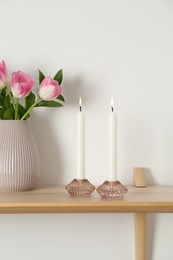 Photo of Elegant holders with burning candles, tulips in vase and other decor on wooden table near white wall
