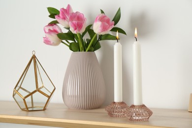 Photo of Elegant holders with burning candles, tulips in vase and other decor on wooden table near white wall