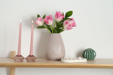 Photo of Elegant holders with burning candles, tulips in vase and other decor on wooden table near white wall