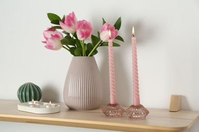 Photo of Elegant holders with burning candles, tulips in vase and other decor on wooden table near white wall
