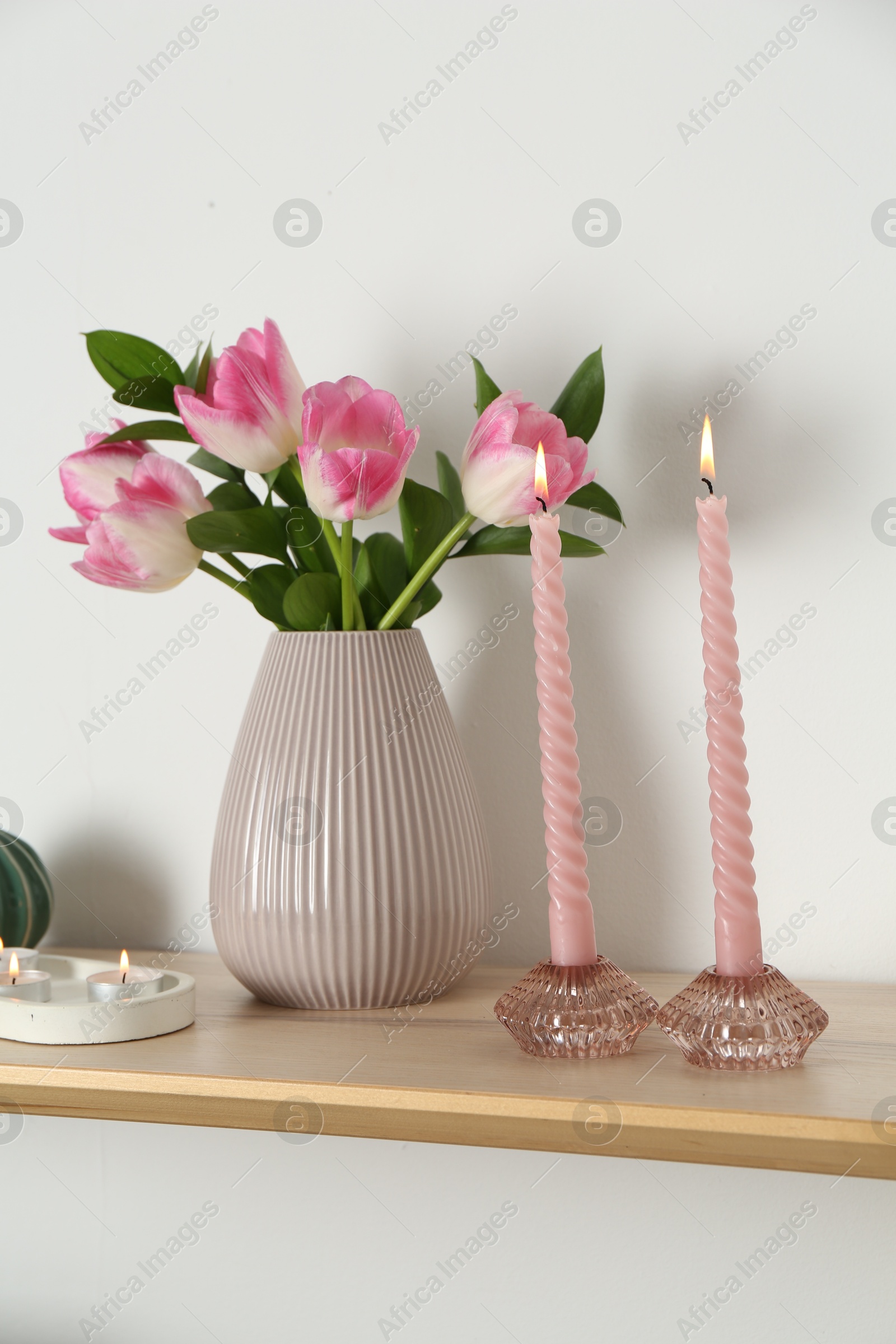 Elegant holders with burning candles, tulips in vase and other decor on wooden table near white wall Photo of Elegant holders with burning candles, tulips in vase and other decor on wooden table near white wall