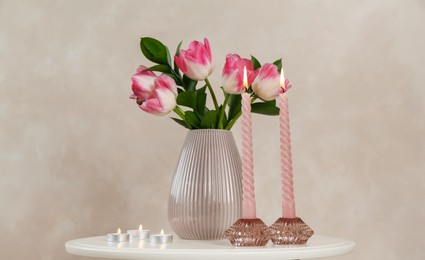 Photo of Elegant holders with burning candles and tulips in vase on white table near beige wall