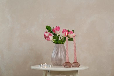 Photo of Elegant holders with burning candles and tulips in vase on white table near beige wall