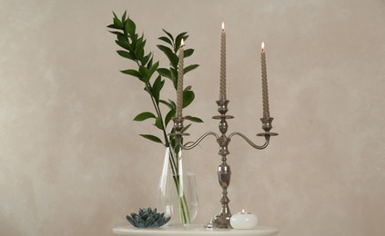 Photo of Elegant candlestick with burning candles, branches in vase and other decor on white table near beige wall