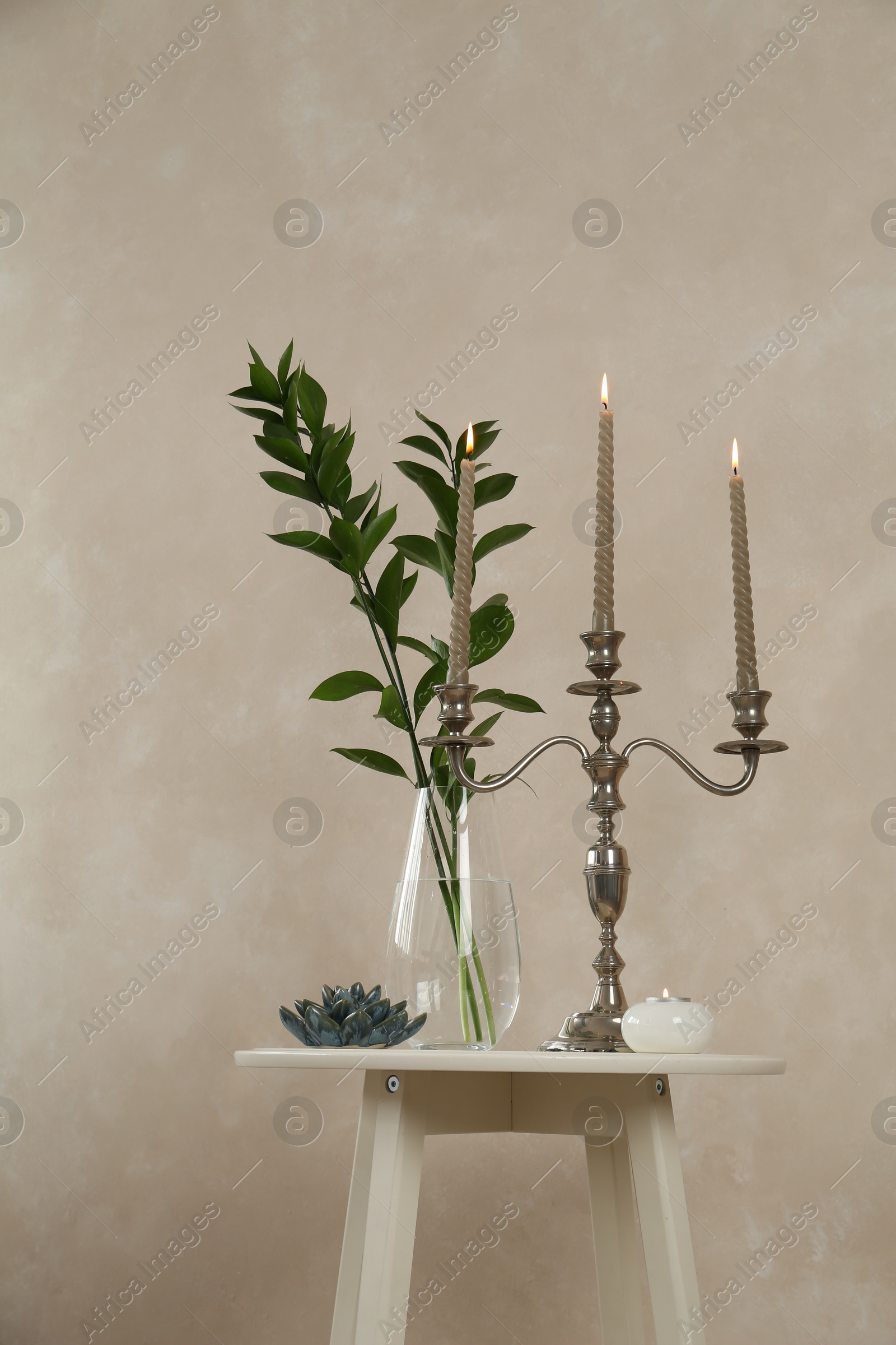 Elegant candlestick with burning candles, branches in vase and other decor on white table near beige wall Photo of Elegant candlestick with burning candles, branches in vase and other decor on white table near beige wall