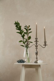 Photo of Elegant candlestick with burning candles, branches in vase and other decor on white table near beige wall