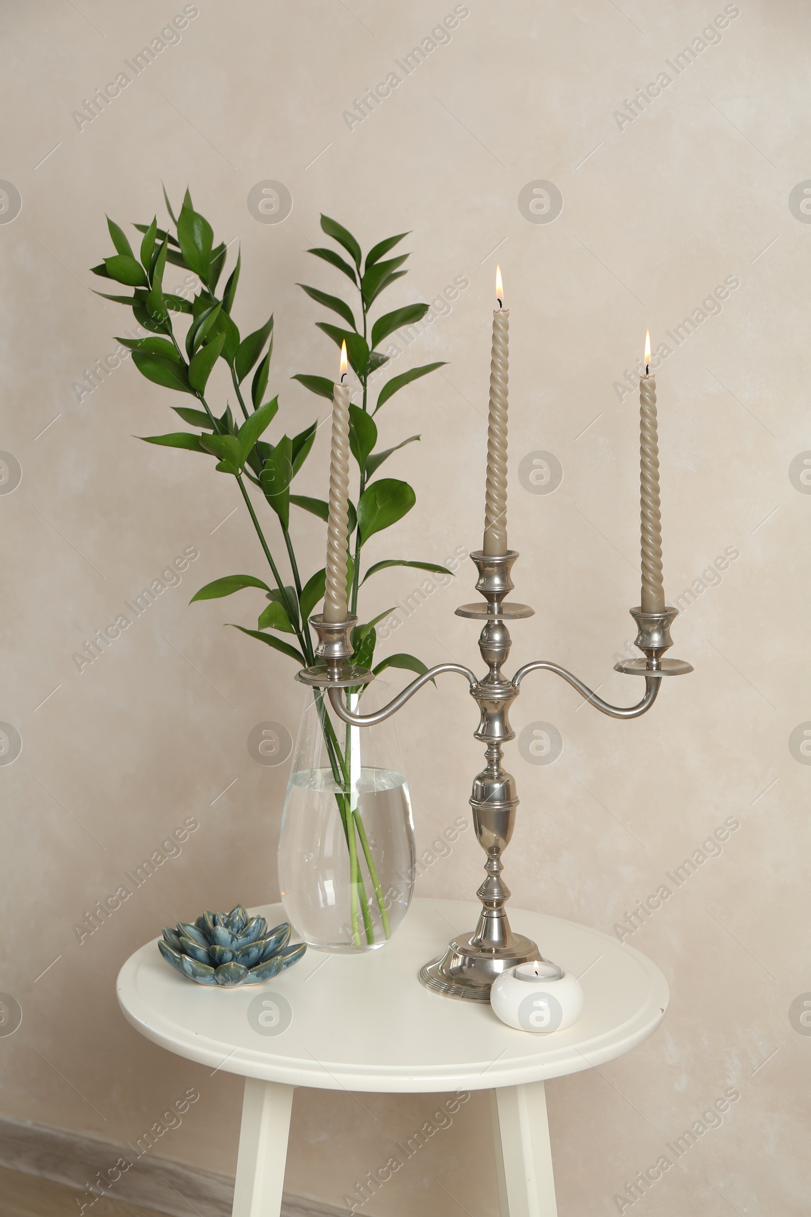Elegant candlestick with burning candles, branches in vase and other decor on white table near beige wall Photo of Elegant candlestick with burning candles, branches in vase and other decor on white table near beige wall