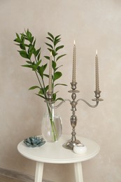 Photo of Elegant candlestick with burning candles, branches in vase and other decor on white table near beige wall