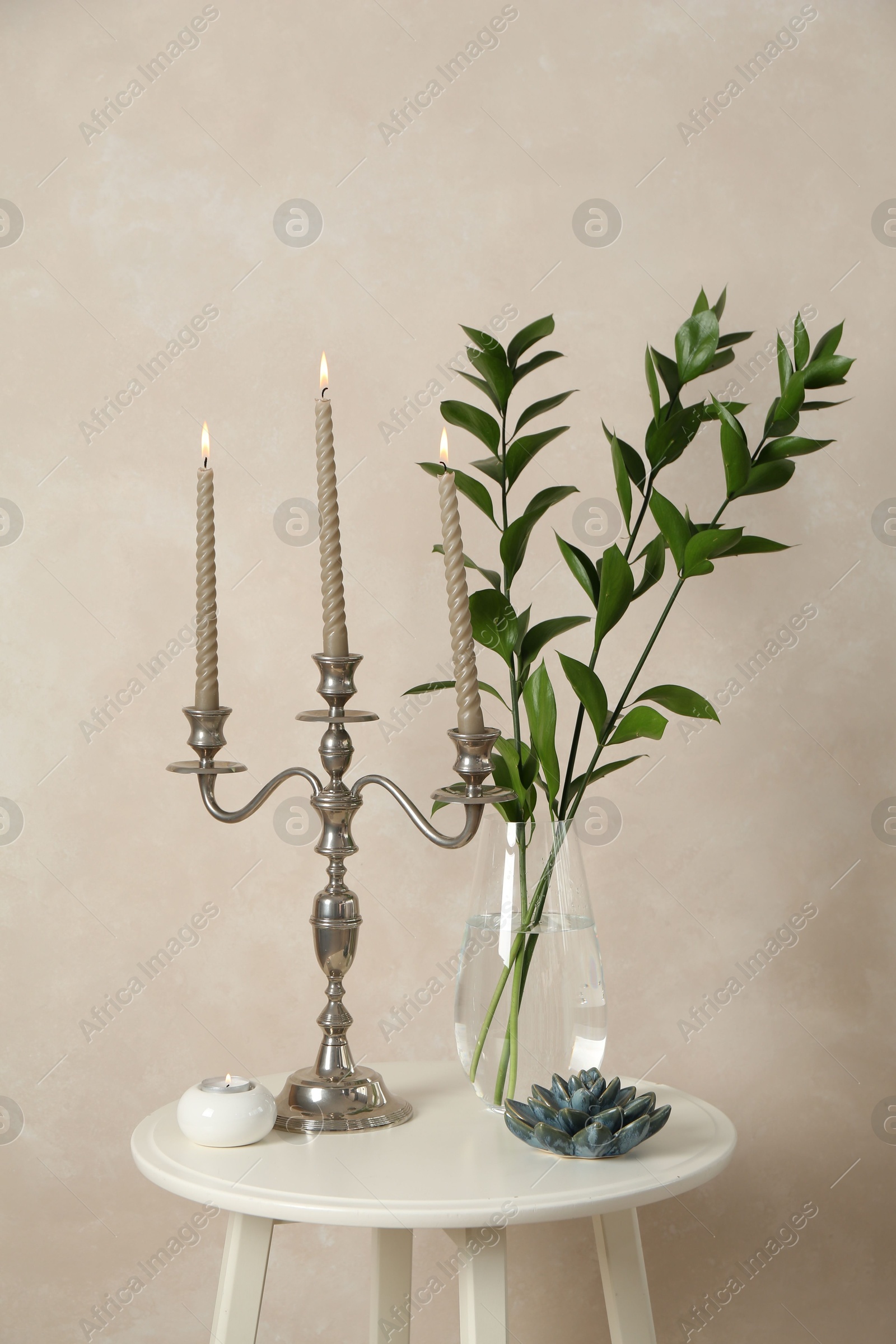 Elegant candlestick with burning candles, branches in vase and other decor on white table near beige wall Photo of Elegant candlestick with burning candles, branches in vase and other decor on white table near beige wall