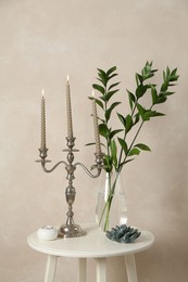 Photo of Elegant candlestick with burning candles, branches in vase and other decor on white table near beige wall
