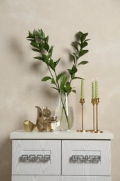 Photo of Elegant candlesticks with burning candles, branches in vase and other decor on dresser near beige wall