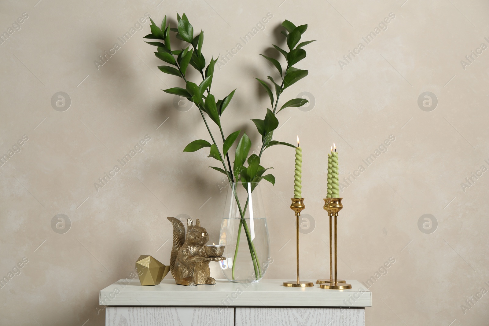 Elegant candlesticks with burning candles, branches in vase and other decor on dresser near beige wall Photo of Elegant candlesticks with burning candles, branches in vase and other decor on dresser near beige wall