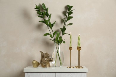 Photo of Elegant candlesticks with burning candles, branches in vase and other decor on dresser near beige wall