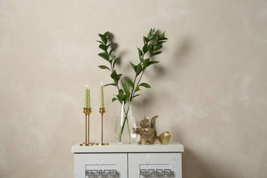 Photo of Elegant candlesticks with burning candles, branches in vase and other decor on dresser near beige wall