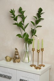 Photo of Elegant candlesticks with burning candles, branches in vase and other decor on dresser near beige wall