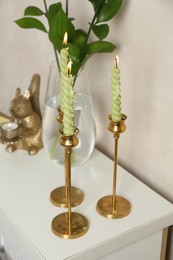 Photo of Elegant candlesticks with burning candles, branches in vase and other decor on dresser near beige wall